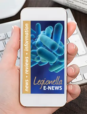 A hand holding a smartphone displaying "Legionella E-News" with a blue bacteria image. The sidebar reads "news, reviews, information." Keyboard blurred in the background.